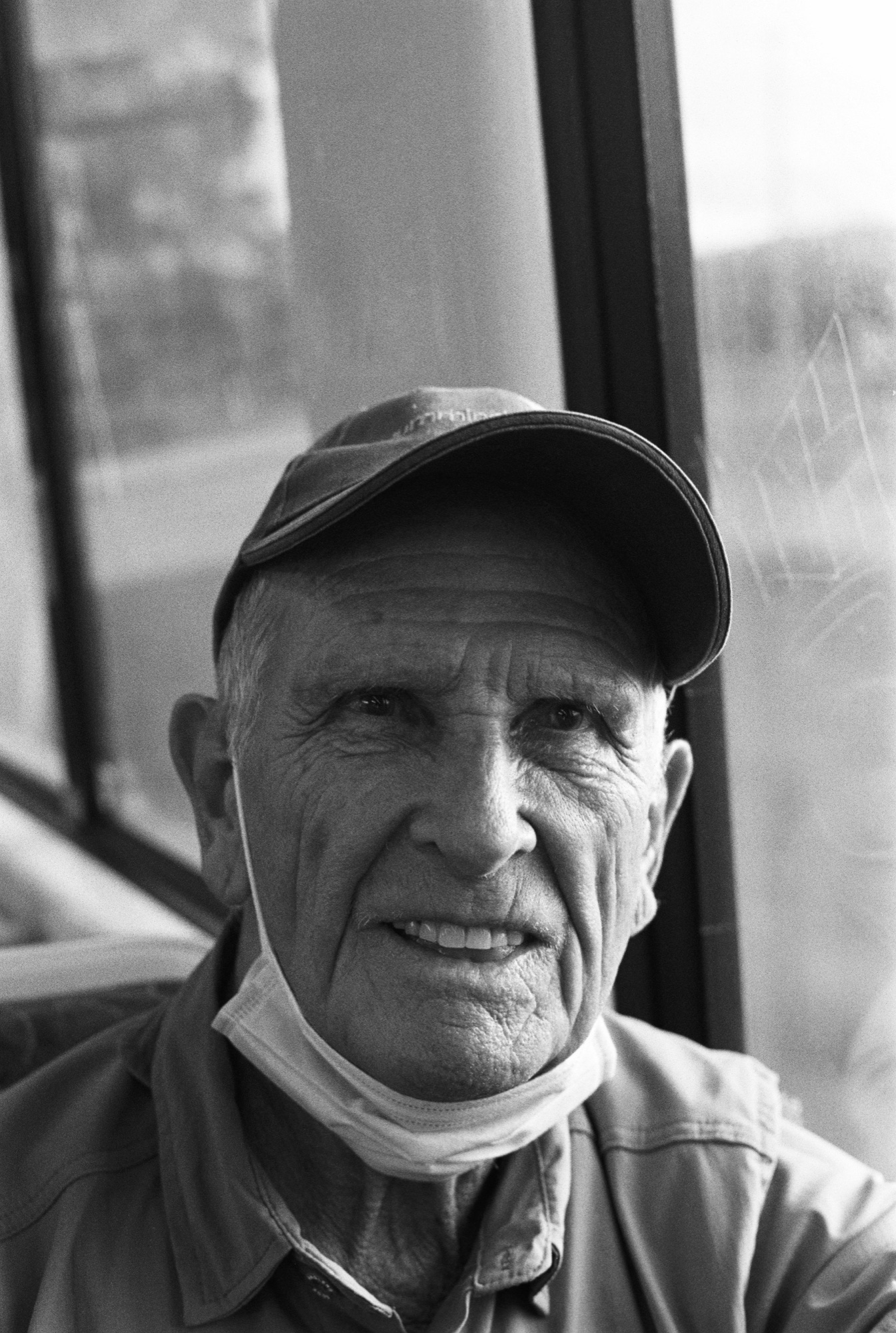 An older Australian man in a cap with a face mask lowered, looking to camera