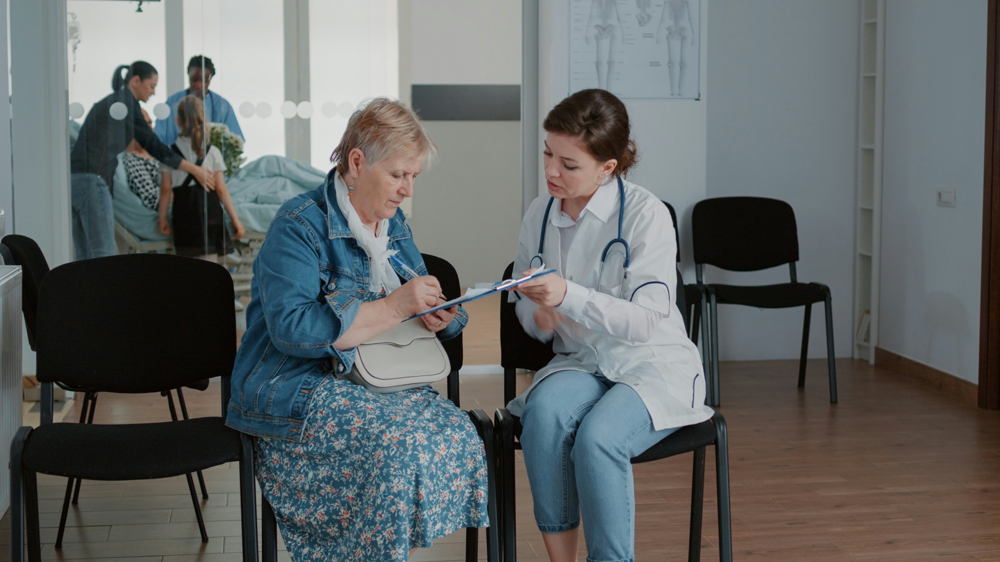 An older patient fills in a form while a clinician sits beside her in a waiting room