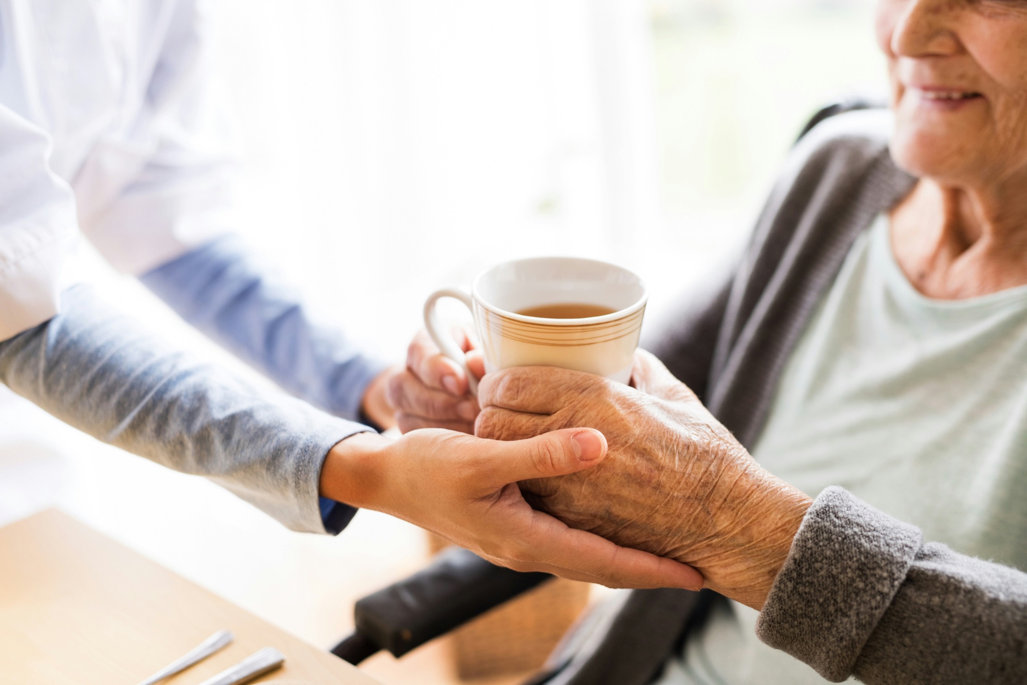 A young carer gently holds an elderly woman's hands around a cup of tea in an aged care home