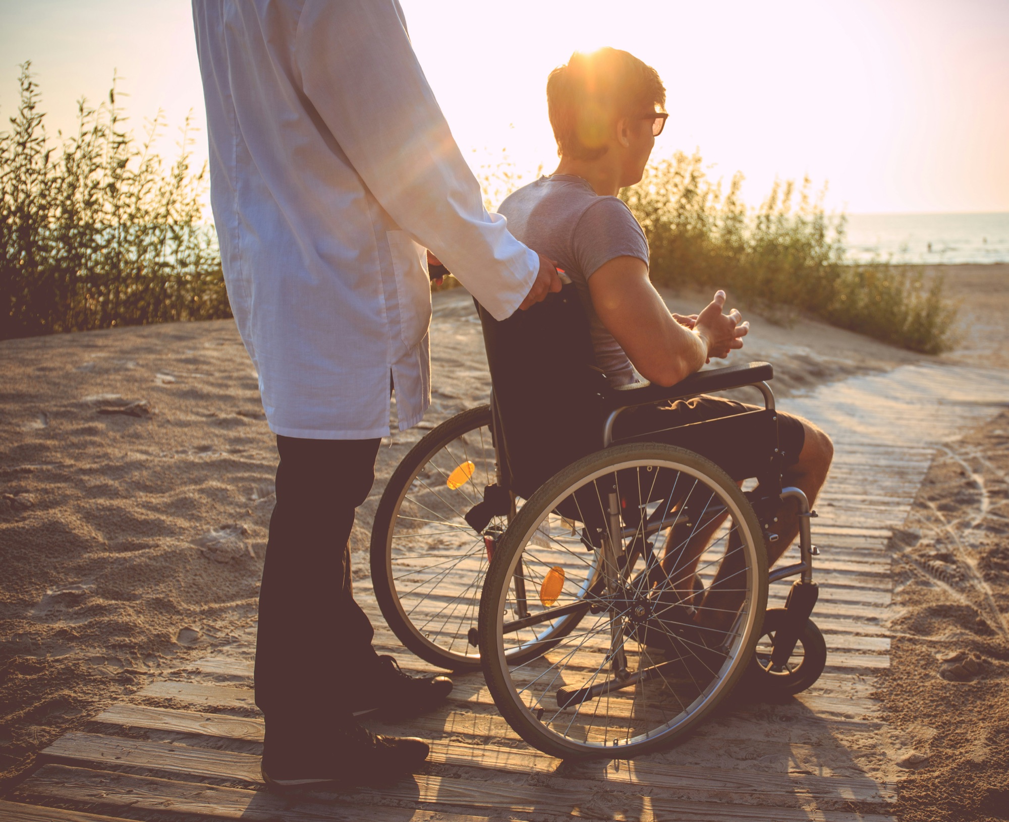 Support worker assisting a man in a wheelchair along a beach boardwalk at sunset